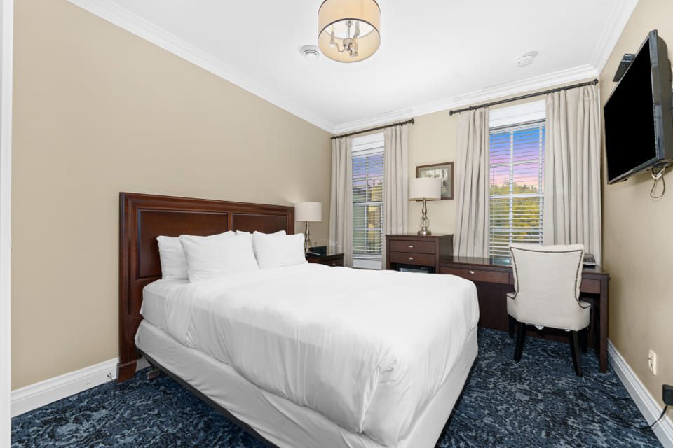 A neatly made bed with white linens in a cozy bedroom featuring beige walls, a dark wooden headboard, a desk with a chair near two tall windows, and a mounted flat-screen TV. 124 on Queen Hotel and Spa | Hotel & Spa Niagara-on-the-lake | 1 (855) 988-4552