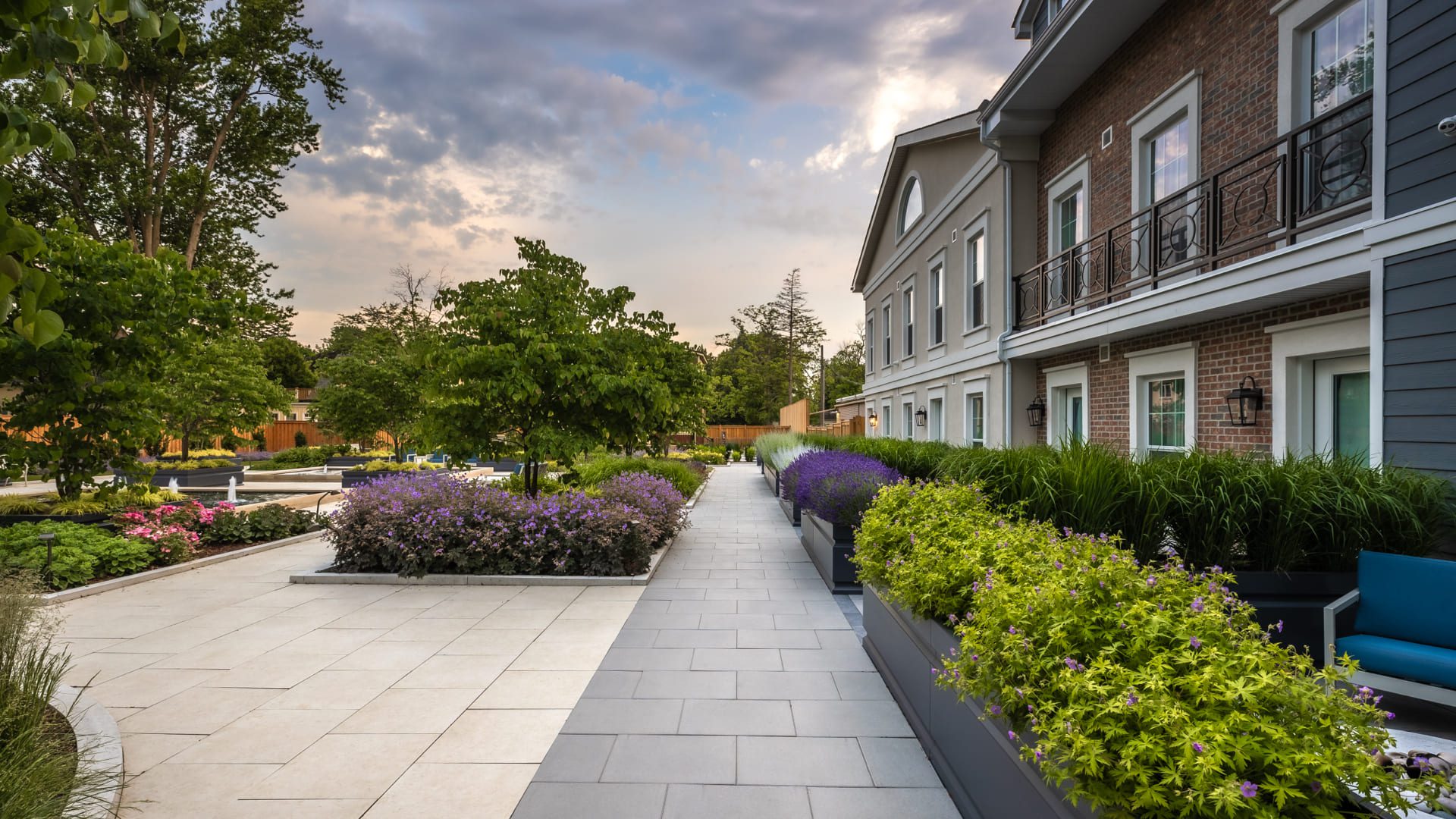 A landscaped walkway lined with lush green plants and purple flowers runs alongside a modern building with balconies, under a partly cloudy sky at sunset. 124 on Queen Hotel and Spa | Hotel & Spa Niagara-on-the-lake | 1 (855) 988-4552