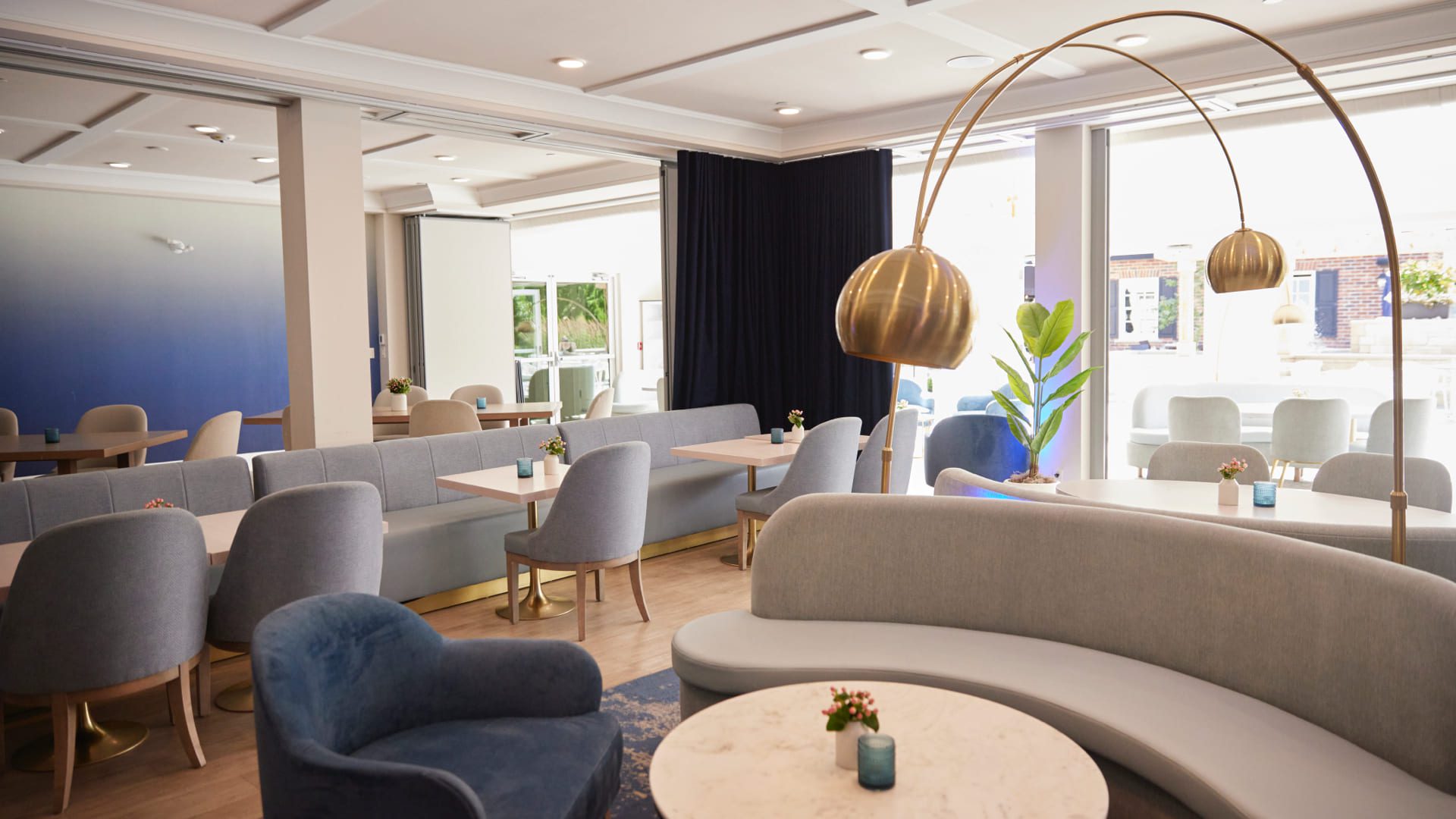 A modern, well-lit lounge with light gray curved sofas, round marble tables, blue chairs, and gold floor lamps—ideal for a winter corporate retreat in Niagara-on-the-Lake. Large windows and navy curtains brighten the spacious, elegant interior. 124 on Queen Hotel and Spa | Hotel & Spa Niagara-on-the-lake | 1 (855) 988-4552