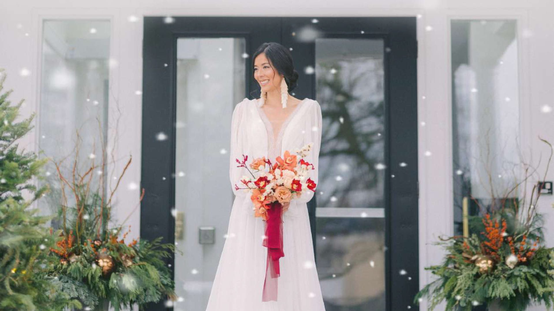 A bride in a white dress stands outside in front of black double doors, holding a bouquet with red and peach flowers. Snow is gently falling, and festive greenery decorates both sides—capturing the magic of winter weddings in Niagara-on-the-Lake. 124 on Queen Hotel and Spa | Hotel & Spa Niagara-on-the-lake | 1 (855) 988-4552