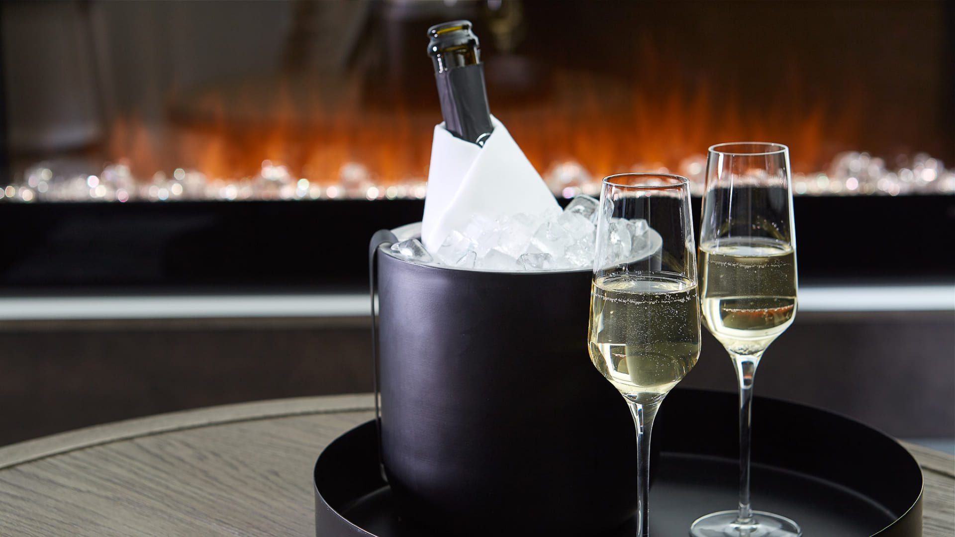 A bottle of champagne chills in an ice bucket beside two flutes on a tray, with a warm fireplace glowing in the background—perfect for winter wine tastings in Niagara-on-the-Lake. 124 on Queen Hotel and Spa | Hotel & Spa Niagara-on-the-lake | 1 (855) 988-4552