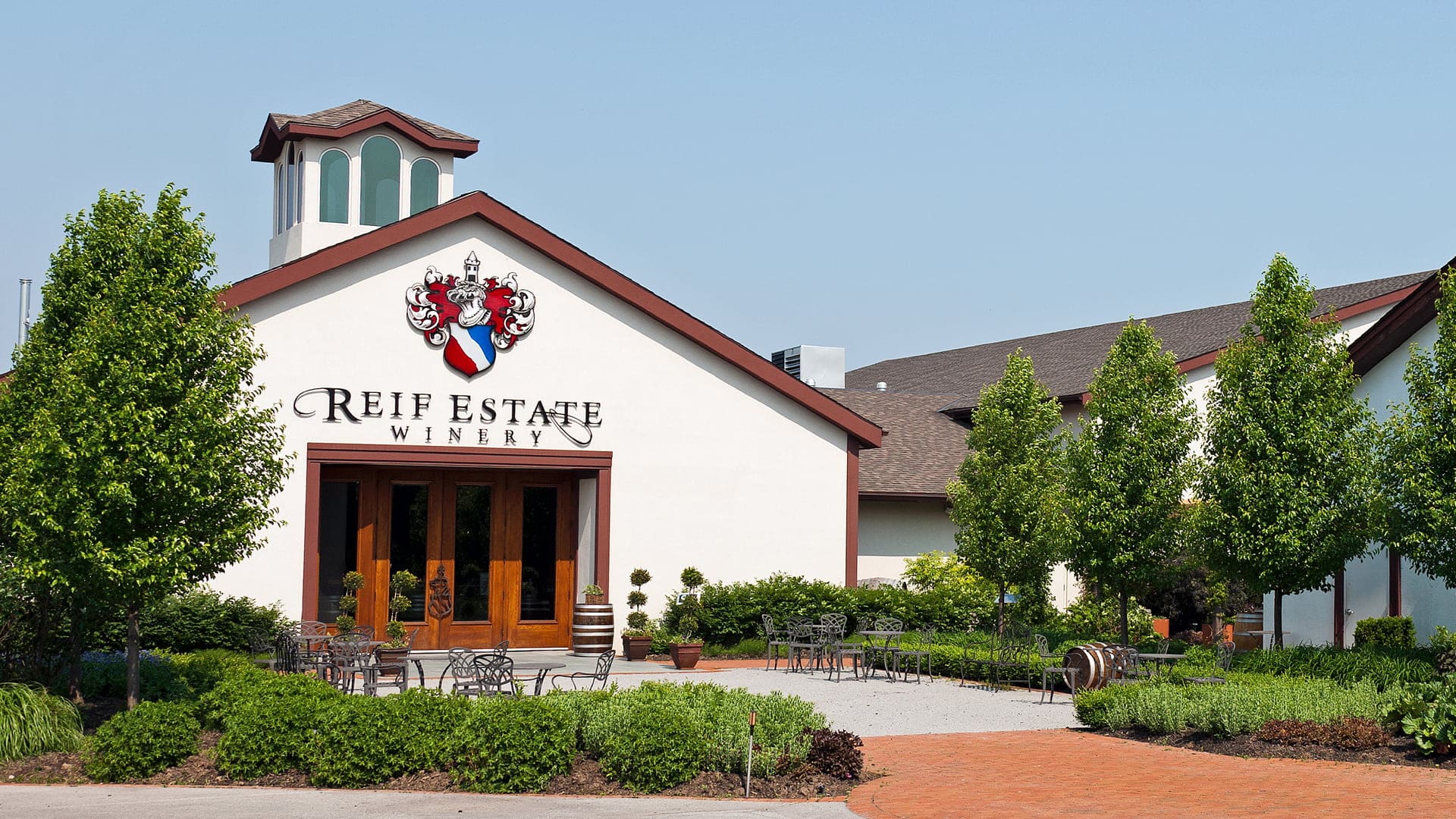 A white building with a peaked roof and a crest sign reading Reif Estate Winery at the entrance, surrounded by green trees, plants, and outdoor patio seating on a sunny day. 124 on Queen Hotel and Spa | Hotel & Spa Niagara-on-the-lake | 1 (855) 988-4552