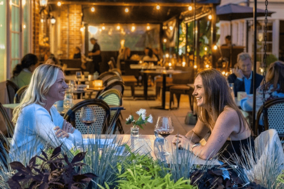 Two women sit at an outdoor restaurant table, smiling and talking. They have wine glasses and a small flower vase on the table. Other diners and warm string lights create a cozy evening atmosphere in the background. 124 on Queen Hotel and Spa | Hotel & Spa Niagara-on-the-lake | 1 (855) 988-4552 Two women sit at an outdoor restaurant table, smiling and talking. They have wine glasses and a small flower vase on the table. Other diners and warm string lights create a cozy evening atmosphere in the background. 124 on Queen Hotel and Spa | Hotel & Spa Niagara-on-the-lake | 1 (855) 988-4552