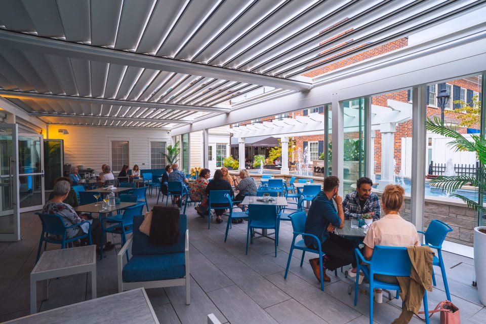 People dine at a covered outdoor patio with blue chairs and tables, surrounded by glass walls and modern decor, enjoying food and drinks in a relaxed, social atmosphere. 124 on Queen Hotel and Spa | Hotel & Spa Niagara-on-the-lake | 1 (855) 988-4552