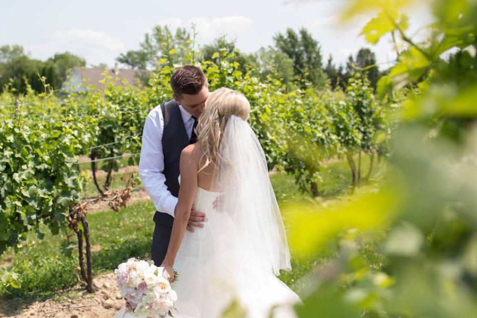 A bride and groom kiss outdoors in a vineyard. The bride wears a white dress and veil, holding a bouquet, while the groom wears a vest and tie. Green vines surround them under a sunny sky. 124 on Queen Hotel and Spa | Hotel & Spa Niagara-on-the-lake | 1 (855) 988-4552