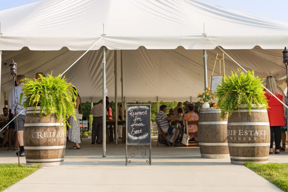 A white event tent with people inside is flanked by large wine barrels topped with ferns. A chalkboard sign in the center reads Reserved for Private Event. The barrels are labeled Reif Estate Winery. 124 on Queen Hotel and Spa | Hotel & Spa Niagara-on-the-lake | 1 (855) 988-4552