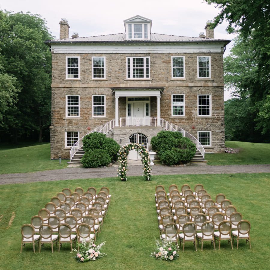 Outdoor wedding ceremony setup on a lawn in front of a large, historic stone building, with rows of chairs facing a floral arch at the base of a staircase leading to the entrance. Lush greenery surrounds the scene. 124 on Queen Hotel and Spa | Hotel & Spa Niagara-on-the-lake | 1 (855) 988-4552