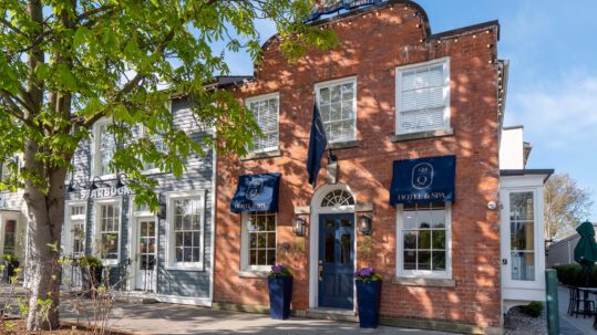 A red-brick boutique hotel in Niagara-on-the-Lake with blue awnings reading “Hotel & Spa,” sits beside a gray building with a Starbucks sign. On this sunny day, a leafy tree casts shadows along the facade and sidewalk. 124 on Queen Hotel and Spa | Hotel & Spa Niagara-on-the-lake | 1 (855) 988-4552