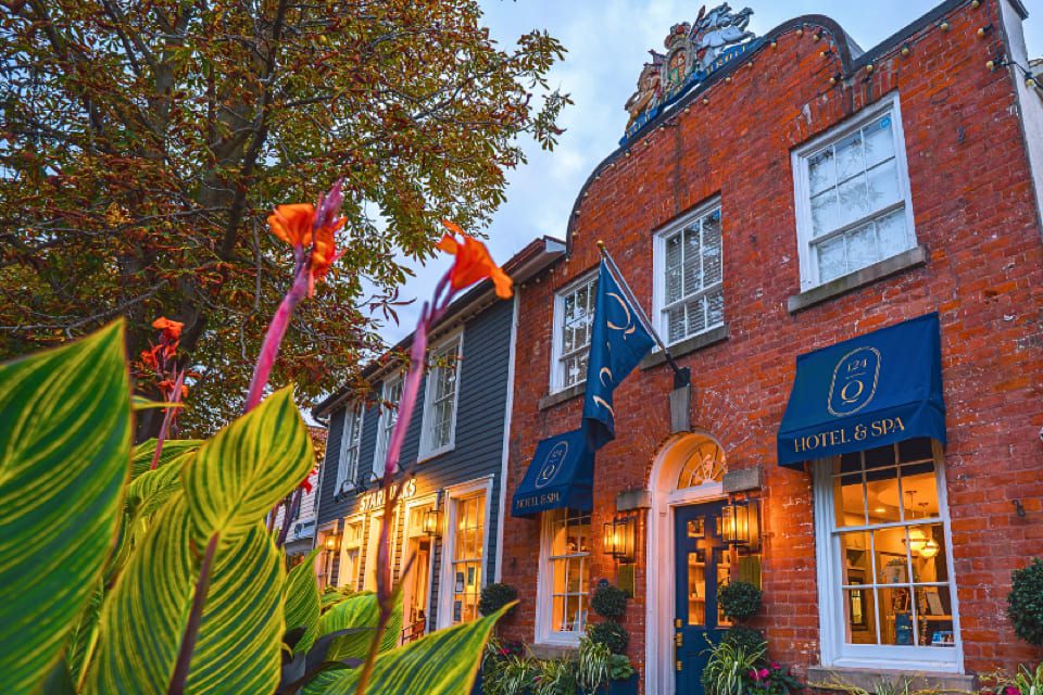 A charming brick building with blue hotel and spa signs, outdoor lanterns, and a decorative crest above the entrance. Colorful flowers and leafy plants are in the foreground, with trees and other quaint buildings nearby. 124 on Queen Hotel and Spa | Hotel & Spa Niagara-on-the-lake | 1 (855) 988-4552