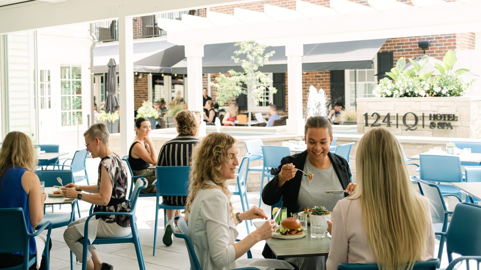 People dining at outdoor patio tables in a bright, modern courtyard. Some are eating and conversing, surrounded by greenery, blue chairs, and a fountain with a sign reading 1241 Q | Hotel & Spa in the background. 124 on Queen Hotel and Spa | Hotel & Spa Niagara-on-the-lake | 1 (855) 988-4552