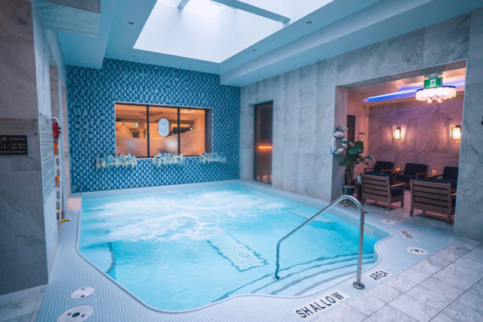 A small indoor pool with clear water, surrounded by white and blue tiles, under a skylight. There is a railing for steps, seating area with chairs, and soft lighting in the background. Signs mark shallow area. 124 on Queen Hotel and Spa | Hotel & Spa Niagara-on-the-lake | 1 (855) 988-4552