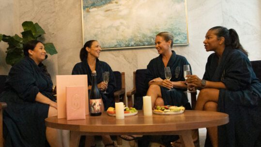Four women in dark robes sit together in a spa lounge, smiling and chatting over champagne glasses—capturing the joy of a girls’ spa getaway in Niagara-on-the-Lake. Candles, snacks, and a pink menu decorate their cozy table. 124 on Queen Hotel and Spa | Hotel & Spa Niagara-on-the-lake | 1 (855) 988-4552