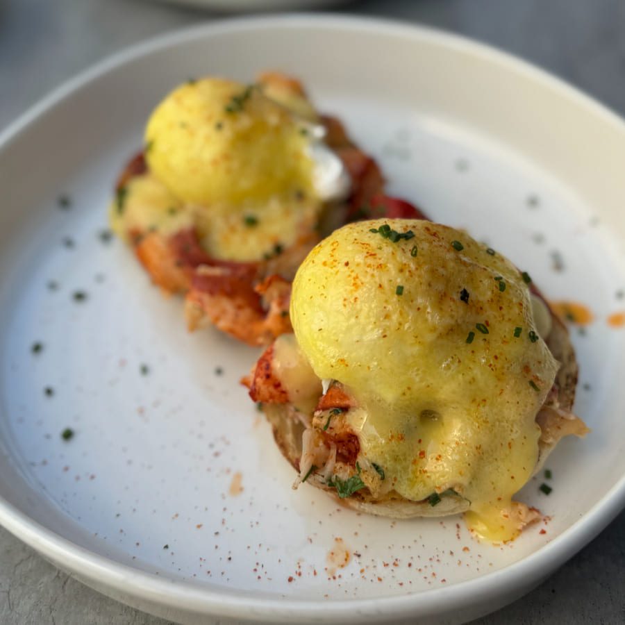 Two eggs Benedict topped with rich hollandaise sauce, garnished with herbs and paprika, served on a white plate. The dish sits on an English muffin with a layer of what appears to be lobster or crab. 124 on Queen Hotel and Spa | Hotel & Spa Niagara-on-the-lake | 1 (855) 988-4552