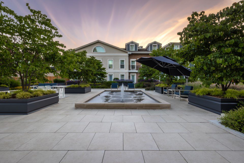 A modern courtyard features a central fountain, large potted plants, seating areas with tables, and a black umbrella. Trees line the space, and houses with dormer windows are visible in the background at sunset. 124 on Queen Hotel and Spa | Hotel & Spa Niagara-on-the-lake | 1 (855) 988-4552