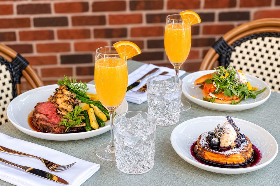 Three plates of gourmet food and two glasses of orange mimosas sit on an outdoor table with a brick wall background; water glasses and neatly arranged napkins and silverware are also present. 124 on Queen Hotel and Spa | Hotel & Spa Niagara-on-the-lake | 1 (855) 988-4552