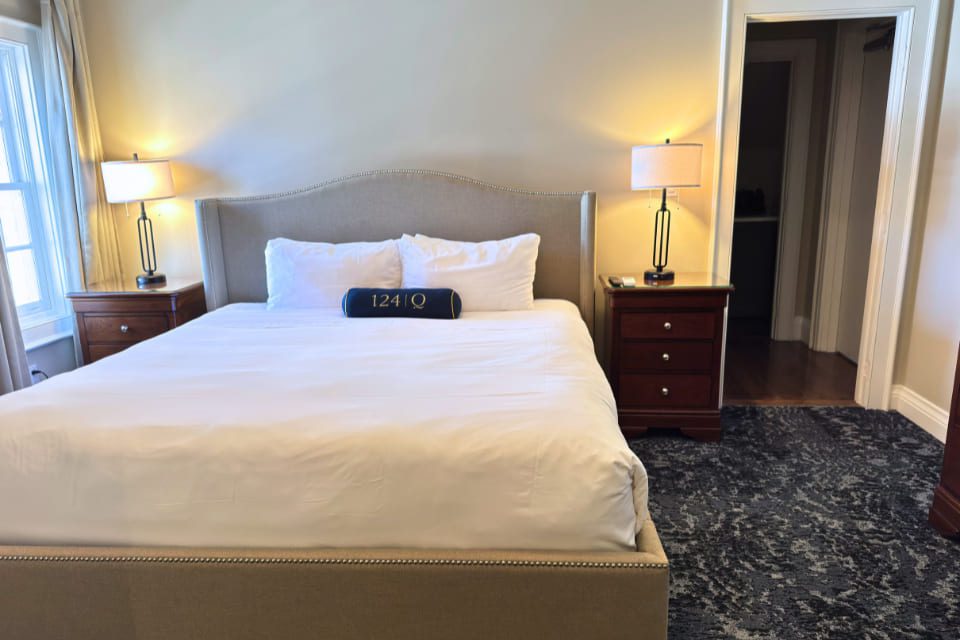 A neatly made bed with white linens and a blue decorative pillow sits next to a nightstand with a lamp in a cozy, well-lit bedroom. An open door reveals a glimpse of a bathroom. 124 on Queen Hotel and Spa | Hotel & Spa Niagara-on-the-lake | 1 (855) 988-4552