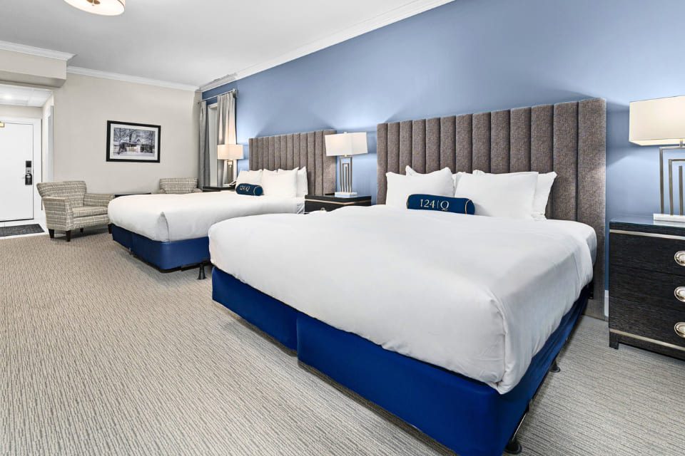 A spacious hotel room with two large beds, blue accent wall, nightstands with lamps, a wall-mounted TV, a desk and chair, large windows with gray curtains, and a potted plant in the corner. 124 on Queen Hotel and Spa | Hotel & Spa Niagara-on-the-lake | 1 (855) 988-4552