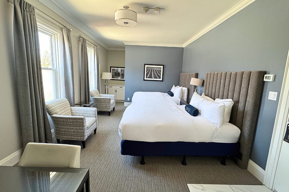 Hotel room with two double beds, gray upholstered headboards, white bedding, a nightstand with a lamp, two patterned armchairs, gray carpet, and framed photos on pale gray walls. Large windows bring in natural light. 124 on Queen Hotel and Spa | Hotel & Spa Niagara-on-the-lake | 1 (855) 988-4552