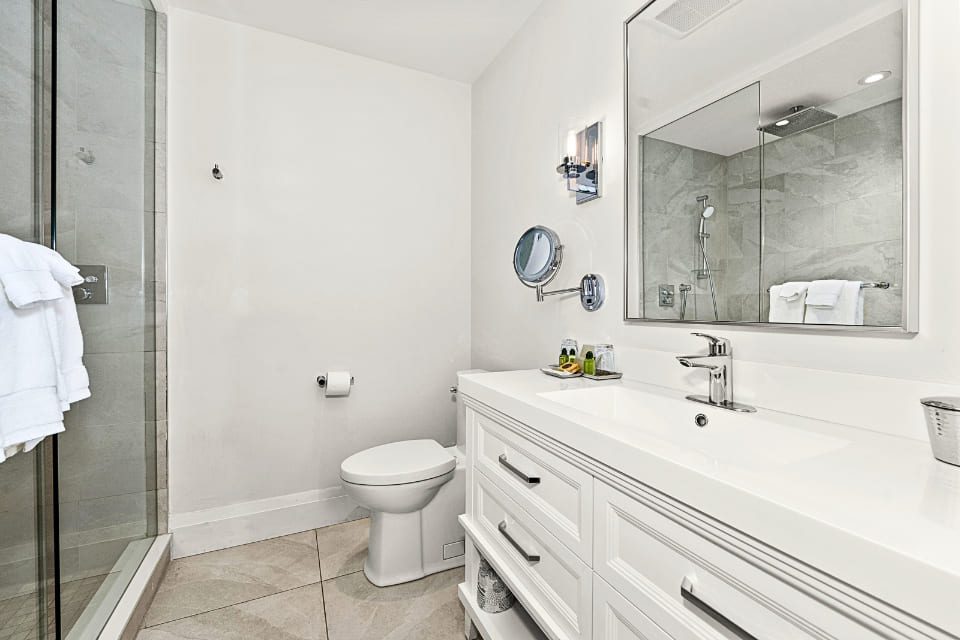 Modern bathroom with a glass shower, white toilet, and double-sink vanity with drawers. Large mirror above the sinks, wall-mounted light fixtures, and neutral tile floor create a clean, bright space. 124 on Queen Hotel and Spa | Hotel & Spa Niagara-on-the-lake | 1 (855) 988-4552