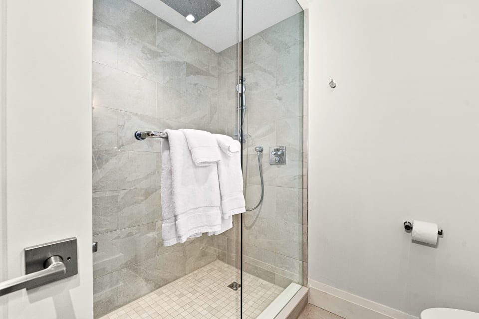 A modern, glass-enclosed shower with light beige tiles and a chrome showerhead. Two white towels hang on a metal bar inside. A toilet paper holder is visible on the wall outside the shower. 124 on Queen Hotel and Spa | Hotel & Spa Niagara-on-the-lake | 1 (855) 988-4552
