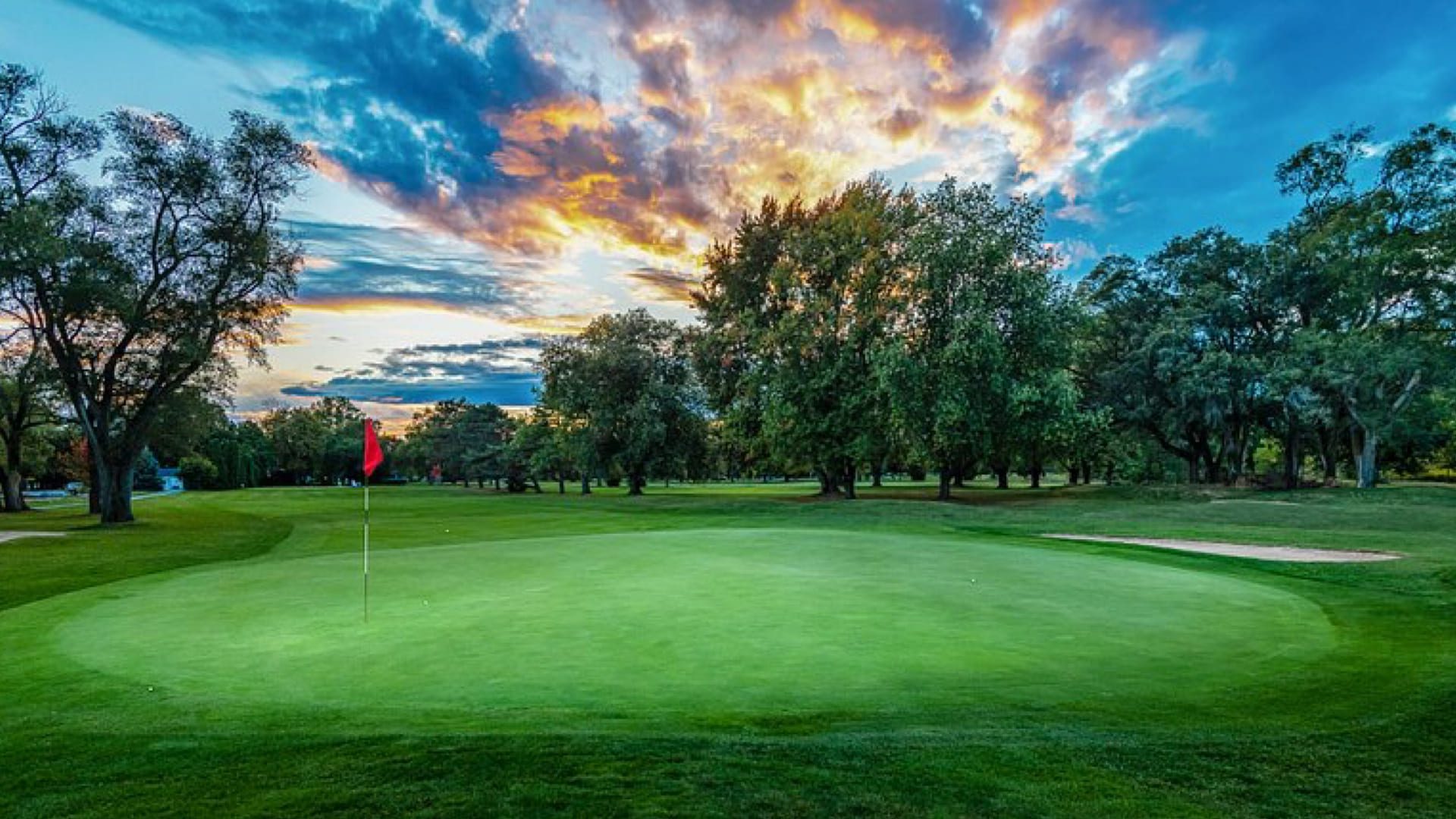 A vibrant sunset illuminates a lush golf course, perfect for a Father’s Day getaway in Niagara-on-the-Lake, with a red flag on the green, surrounded by trees and a sand bunker under a dramatic, colorful sky. 124 on Queen Hotel and Spa | Hotel & Spa Niagara-on-the-lake | 1 (855) 988-4552