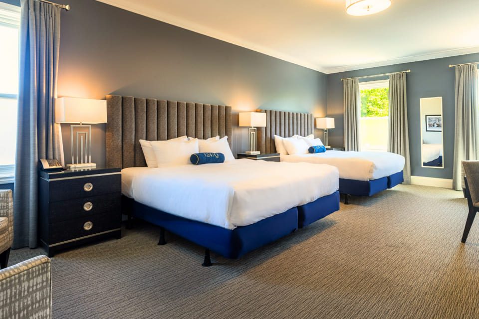 A modern hotel room with two large beds, gray upholstered headboards, blue bases, white bedding, blue accent pillows, nightstands with lamps, large windows with gray curtains, and a carpeted floor. 124 on Queen Hotel and Spa | Hotel & Spa Niagara-on-the-lake | 1 (855) 988-4552