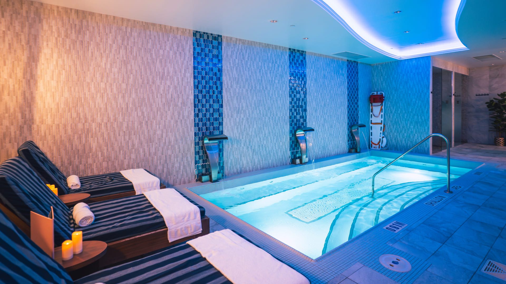 Indoor spa area with a small illuminated pool, striped lounge chairs with rolled towels, and candles. The modern decor and blue ambient lighting make it perfect for a relaxing Father’s Day getaway in Niagara-on-the-Lake. 124 on Queen Hotel and Spa | Hotel & Spa Niagara-on-the-lake | 1 (855) 988-4552