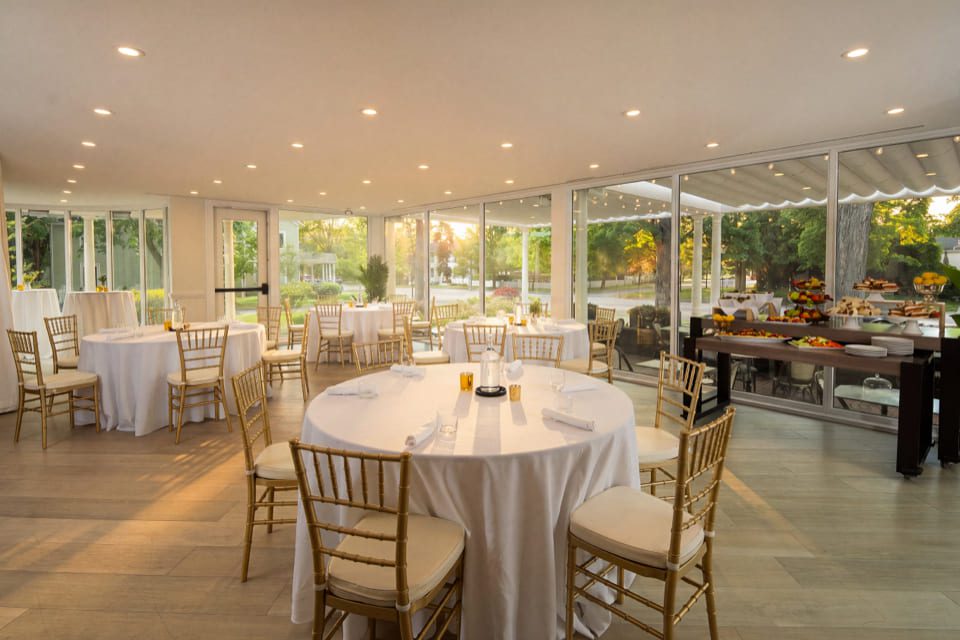 Elegant dining room with round tables covered in white cloths, gold chairs, and large windows showing a garden at sunset. Place settings and candles are arranged on the tables. 124 on Queen Hotel and Spa | Hotel & Spa Niagara-on-the-lake | 1 (855) 988-4552