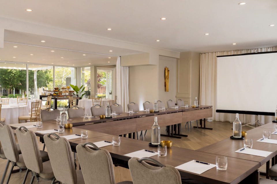 Modern conference room with U-shaped tables, beige chairs, notepads, pens, water bottles, and glasses. A projector screen is at the front, and large windows let in natural light. 124 on Queen Hotel and Spa | Hotel & Spa Niagara-on-the-lake | 1 (855) 988-4552