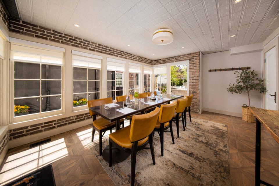 A dining room with a long table set for eight, yellow chairs, brick and white walls, large windows, and a plant in the corner. Natural light fills the space, creating a cozy, inviting atmosphere. 124 on Queen Hotel and Spa | Hotel & Spa Niagara-on-the-lake | 1 (855) 988-4552