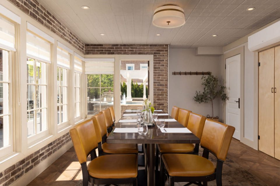 A dining room with a long table set for eight, yellow chairs, brick and white walls, large windows, and a plant in the corner. Natural light fills the space, creating a cozy, inviting atmosphere. 124 on Queen Hotel and Spa | Hotel & Spa Niagara-on-the-lake | 1 (855) 988-4552