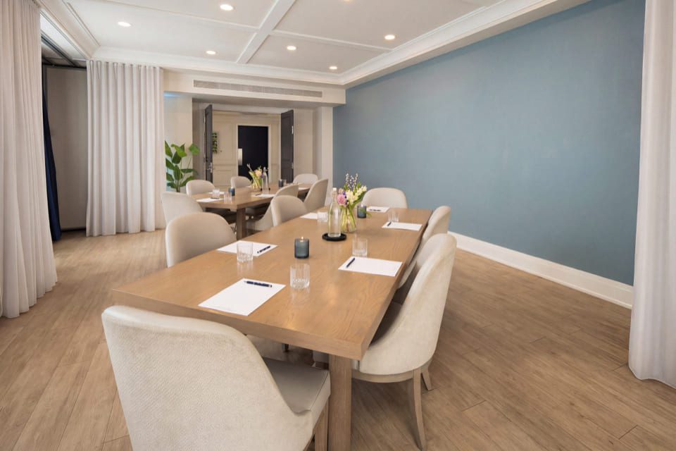 A modern meeting room with a wooden table, beige chairs, notepads, pens, glasses of water, and vases with flowers. The room has light wood flooring, blue and white walls, and ceiling lights. 124 on Queen Hotel and Spa | Hotel & Spa Niagara-on-the-lake | 1 (855) 988-4552