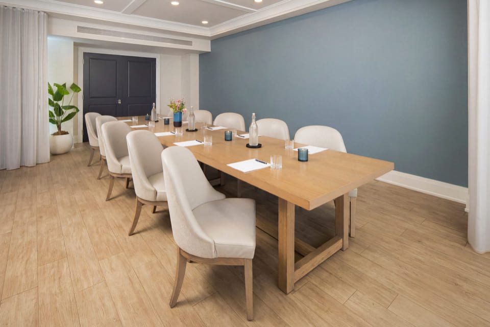 A modern meeting room with a wooden table, beige chairs, notepads, pens, glasses of water, and vases with flowers. The room has light wood flooring, blue and white walls, and ceiling lights. 124 on Queen Hotel and Spa | Hotel & Spa Niagara-on-the-lake | 1 (855) 988-4552