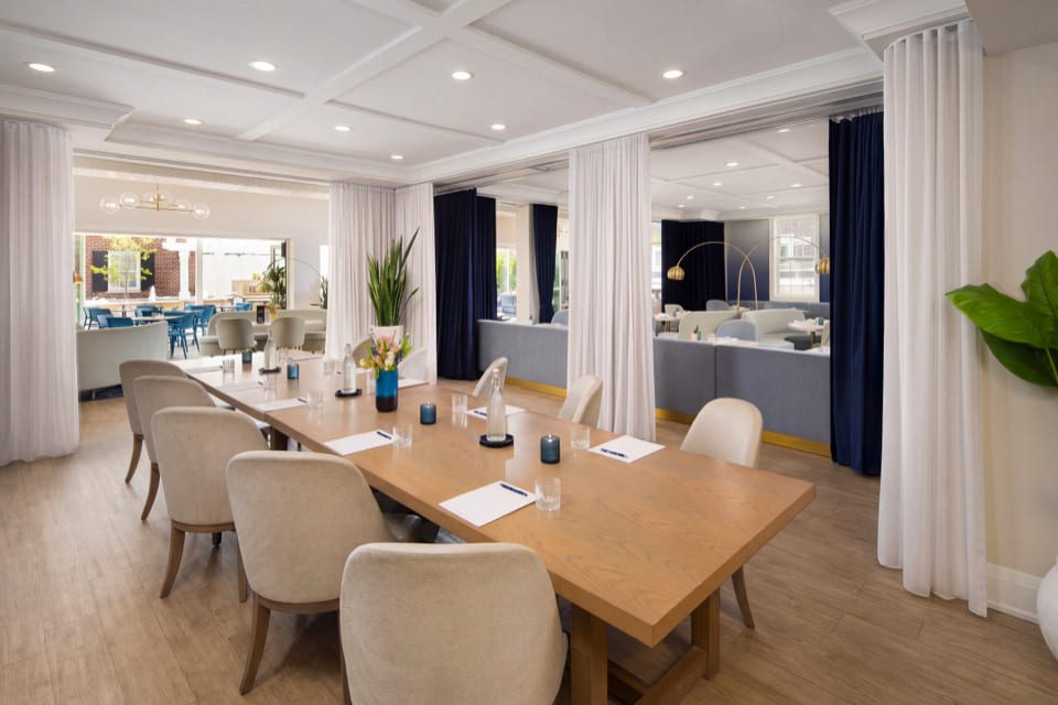 A conference room with a long wooden table, beige chairs, notepads with pens, water glasses, vases with flowers, and curtains separating the space. Bright lighting and windows showing an outdoor area. 124 on Queen Hotel and Spa | Hotel & Spa Niagara-on-the-lake | 1 (855) 988-4552