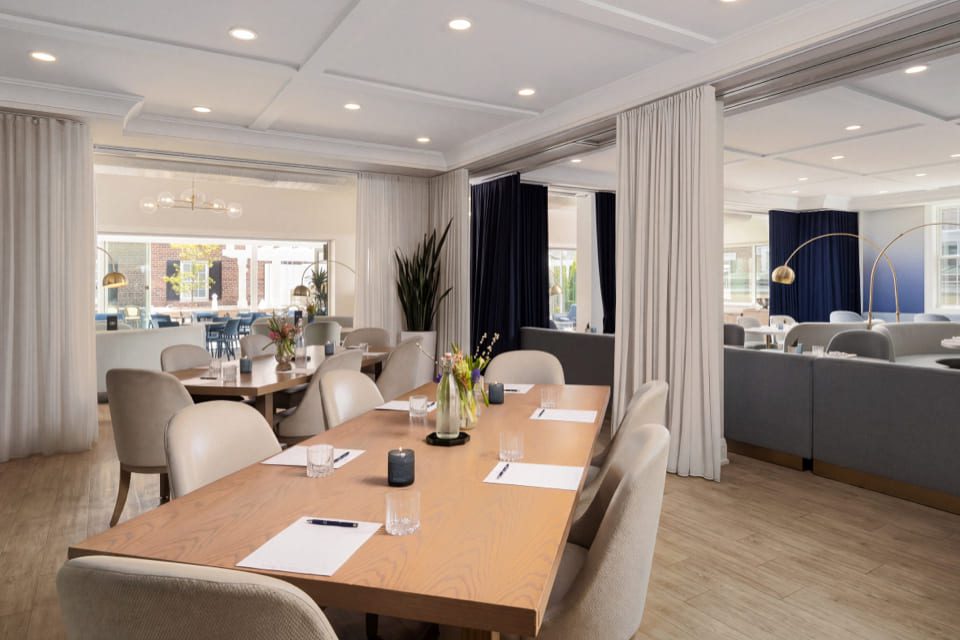 A conference room with a long wooden table, beige chairs, notepads with pens, water glasses, vases with flowers, and curtains separating the space. Bright lighting and windows showing an outdoor area. 124 on Queen Hotel and Spa | Hotel & Spa Niagara-on-the-lake | 1 (855) 988-4552