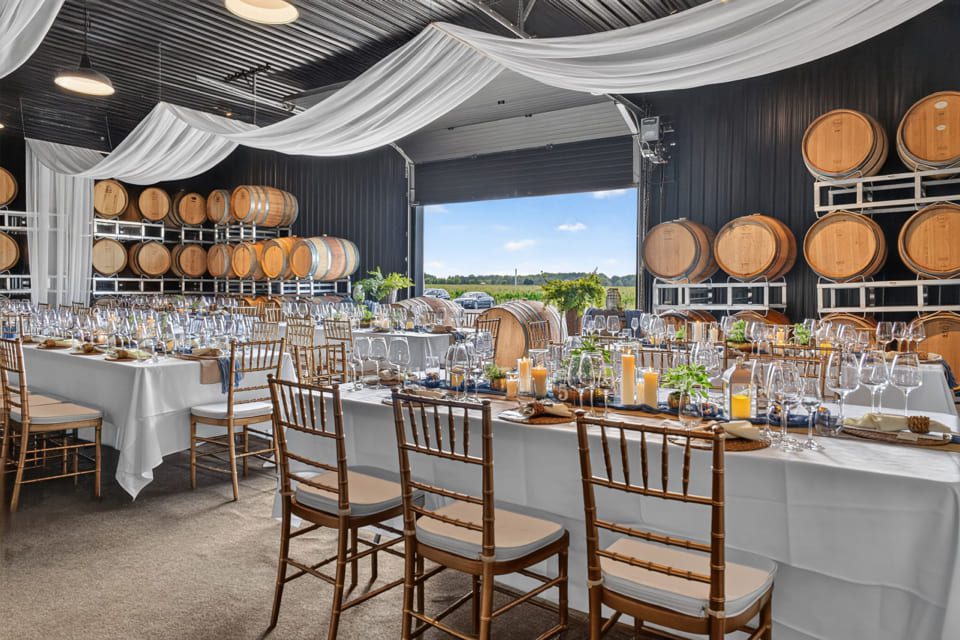 Elegant winery event space with long tables set for dining, gold chairs, white drapes on the ceiling, wine barrels lining the walls, and candles as centerpieces; large open door reveals outdoor scenery. 124 on Queen Hotel and Spa | Hotel & Spa Niagara-on-the-lake | 1 (855) 988-4552