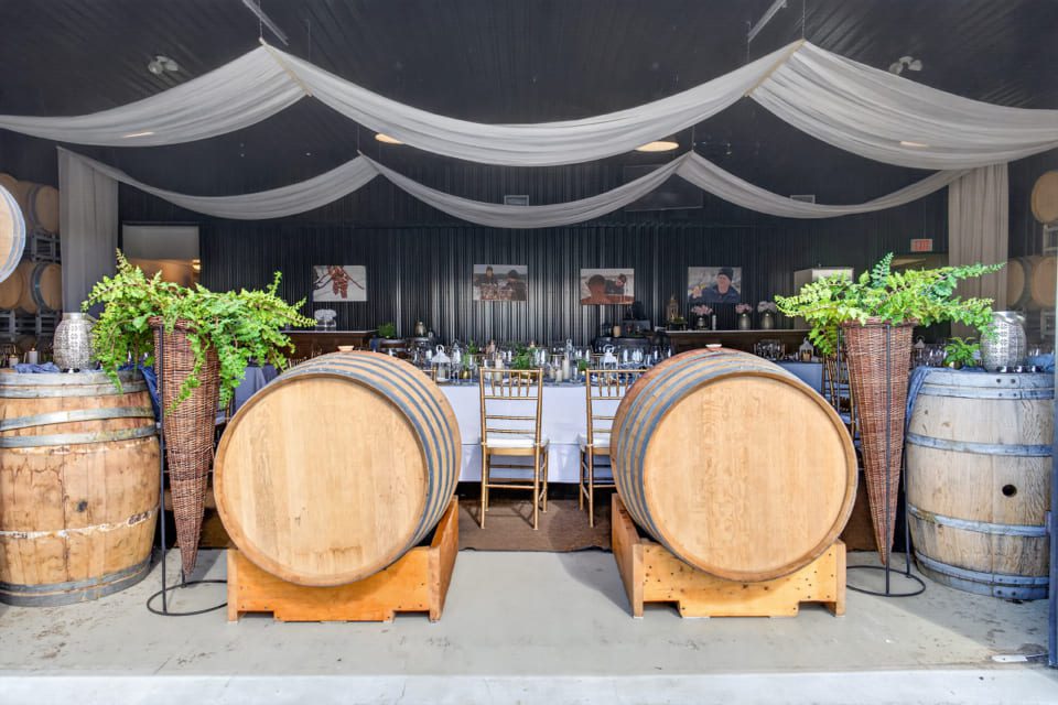 Elegant winery event setup with tables and chairs inside, two large wooden barrels in front, draped white fabric on the ceiling, greenery arrangements, and wine barrels along the walls. 124 on Queen Hotel and Spa | Hotel & Spa Niagara-on-the-lake | 1 (855) 988-4552