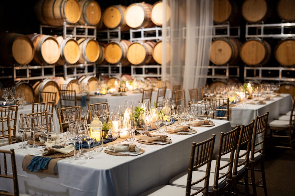 Elegant dinner setup in a winery, with tables set with candles, wine glasses, and plates, surrounded by wooden barrels stacked on racks in the background. Gold chairs are neatly arranged around the tables. 124 on Queen Hotel and Spa | Hotel & Spa Niagara-on-the-lake | 1 (855) 988-4552