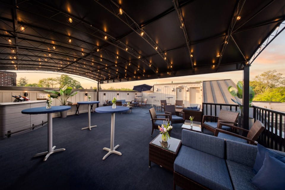 Rooftop patio at sunset with wicker sofas and chairs, blue cushions, string lights, potted plants, and small tables with flowers, creating a cozy, inviting atmosphere. 124 on Queen Hotel and Spa | Hotel & Spa Niagara-on-the-lake | 1 (855) 988-4552