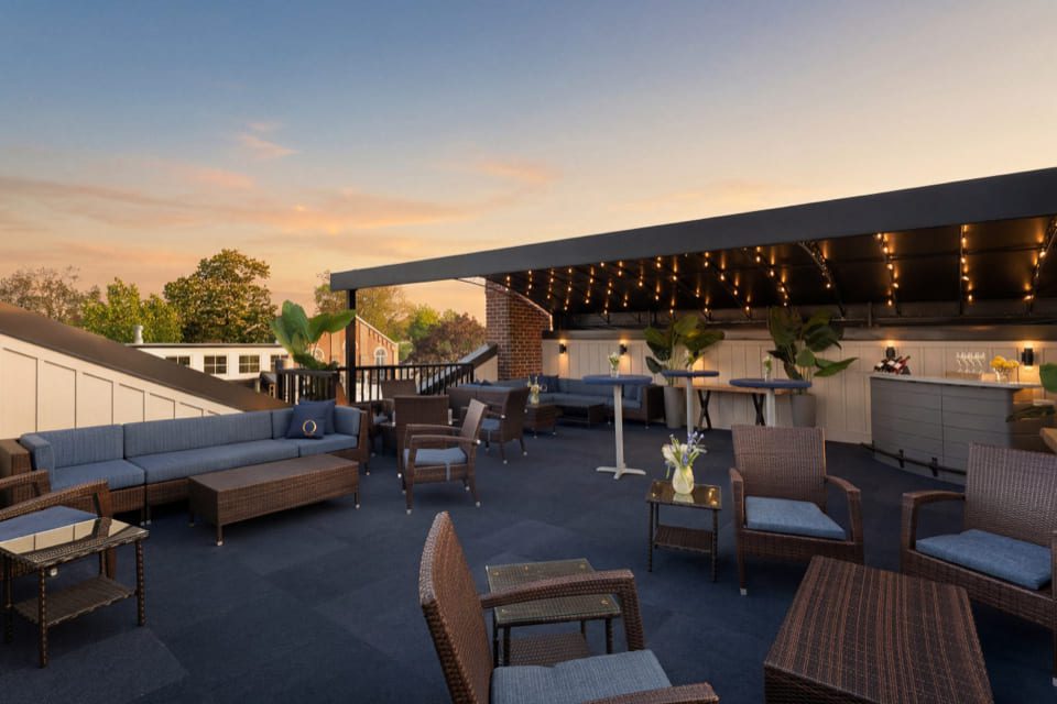 Rooftop patio at sunset with wicker sofas and chairs, blue cushions, string lights, potted plants, and small tables with flowers, creating a cozy, inviting atmosphere. 124 on Queen Hotel and Spa | Hotel & Spa Niagara-on-the-lake | 1 (855) 988-4552