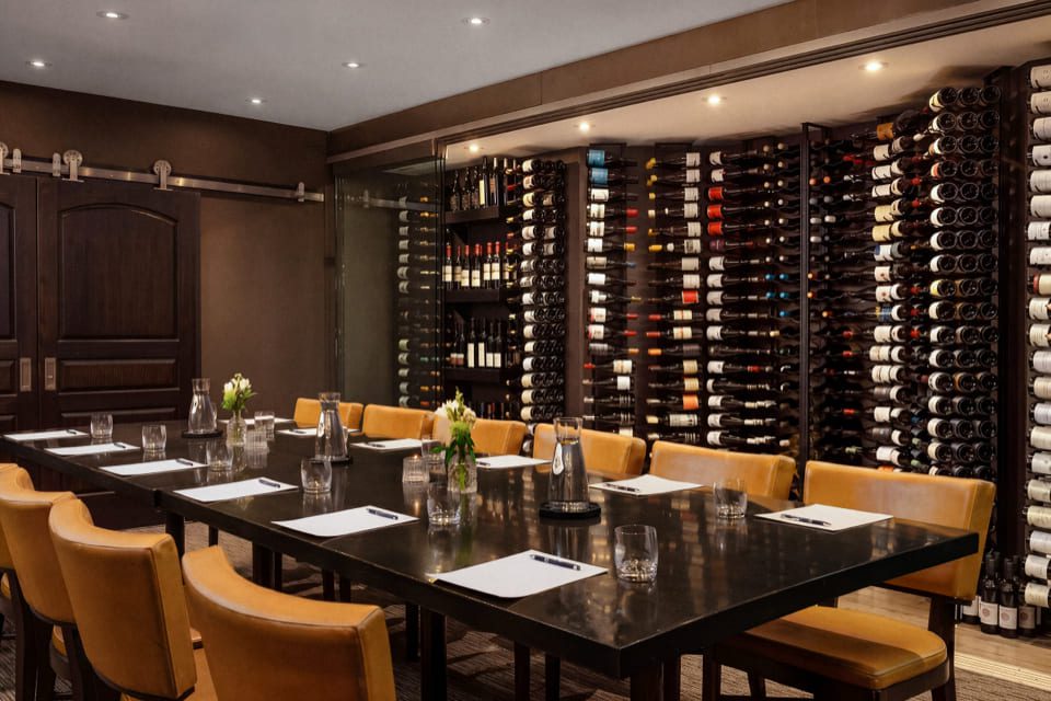 A dining table with yellow chairs is set with glassware and napkins, in front of a wall filled with wine bottles in a dimly lit, elegant restaurant. 124 on Queen Hotel and Spa | Hotel & Spa Niagara-on-the-lake | 1 (855) 988-4552
