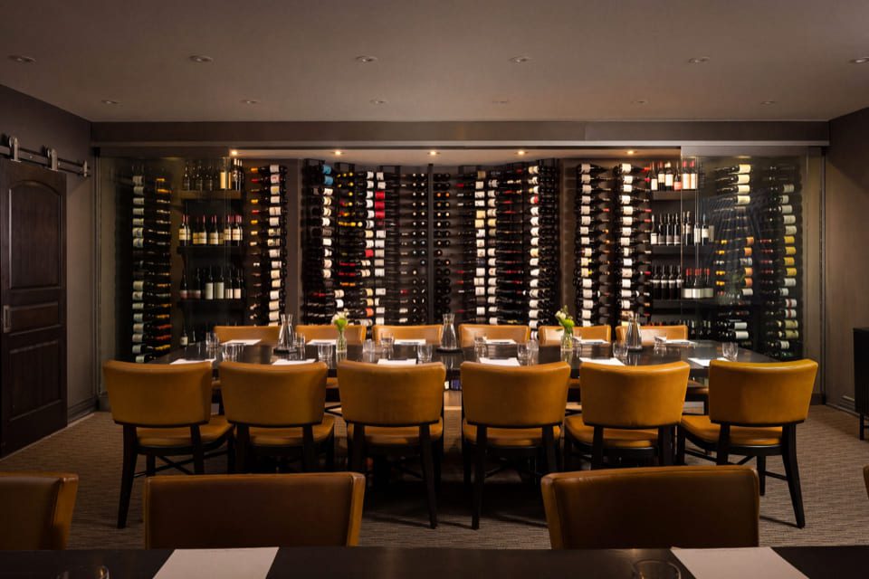 A dining table with yellow chairs is set with glassware and napkins, in front of a wall filled with wine bottles in a dimly lit, elegant restaurant. 124 on Queen Hotel and Spa | Hotel & Spa Niagara-on-the-lake | 1 (855) 988-4552