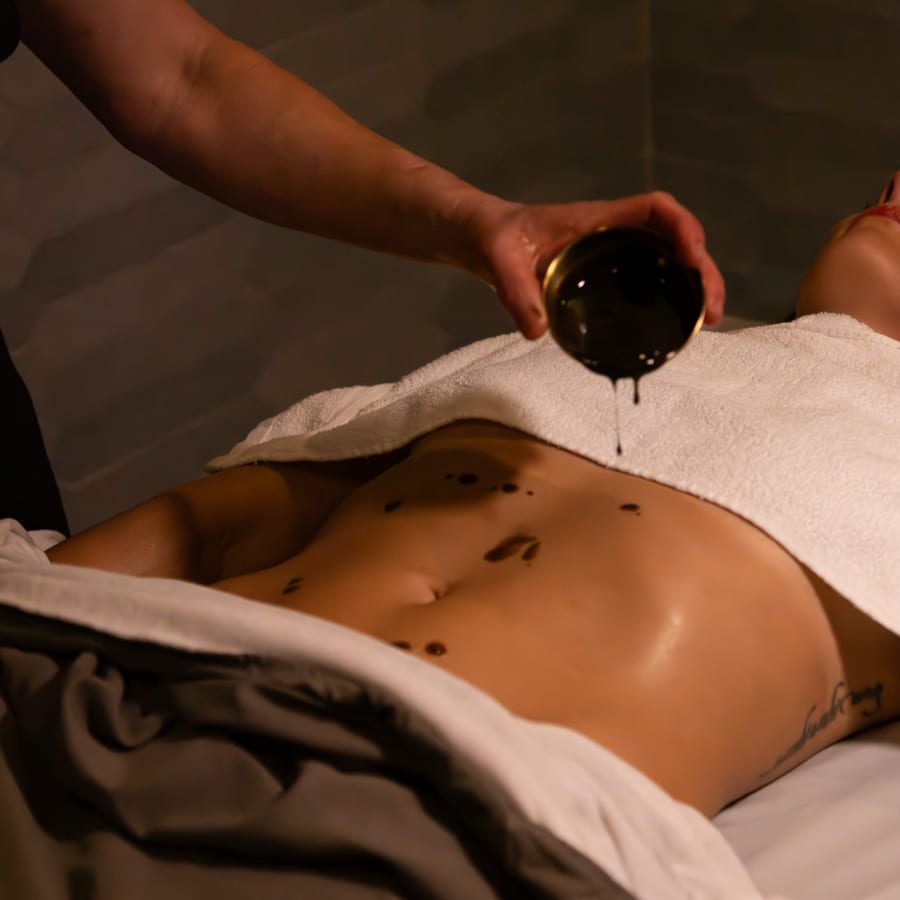 A person lying down with a towel covering their chest while another person pours oil onto their stomach, preparing for a massage. 124 on Queen Hotel and Spa | Hotel & Spa Niagara-on-the-lake | 1 (855) 988-4552