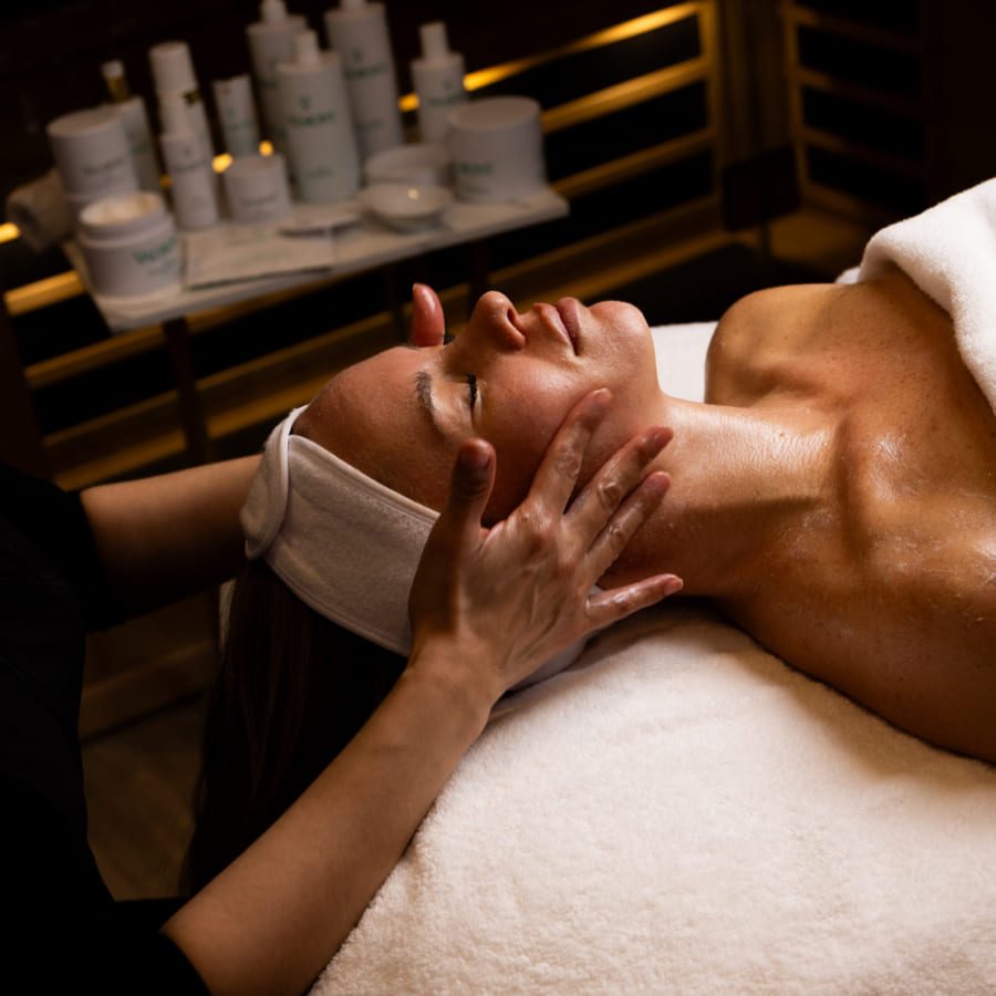 A person lies on a spa bed with a towel covering their body, receiving a facial massage. Skincare products are visible in the background under warm lighting. 124 on Queen Hotel and Spa | Hotel & Spa Niagara-on-the-lake | 1 (855) 988-4552