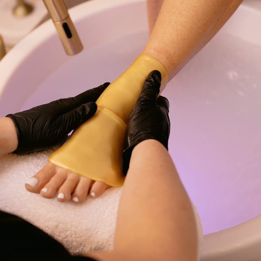 A person wearing black gloves applies a golden foot mask to another persons foot during a spa treatment. The foot rests on a white towel above a basin filled with water. 124 on Queen Hotel and Spa | Hotel & Spa Niagara-on-the-lake | 1 (855) 988-4552